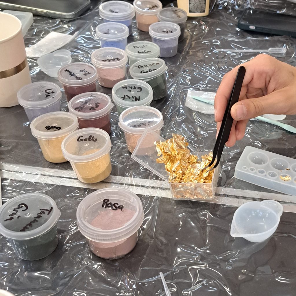 Bild eines KIndergeburtstag Schmuckworkshop von BellaEstellaDesign, der am 2025-09-21 stattgefunden hat. Abgebildet ist die Hand eines Kindes, das Metallflocken mit der Pinzette aufnimmt, daneben steht Farbpulver auf dem Tisch.